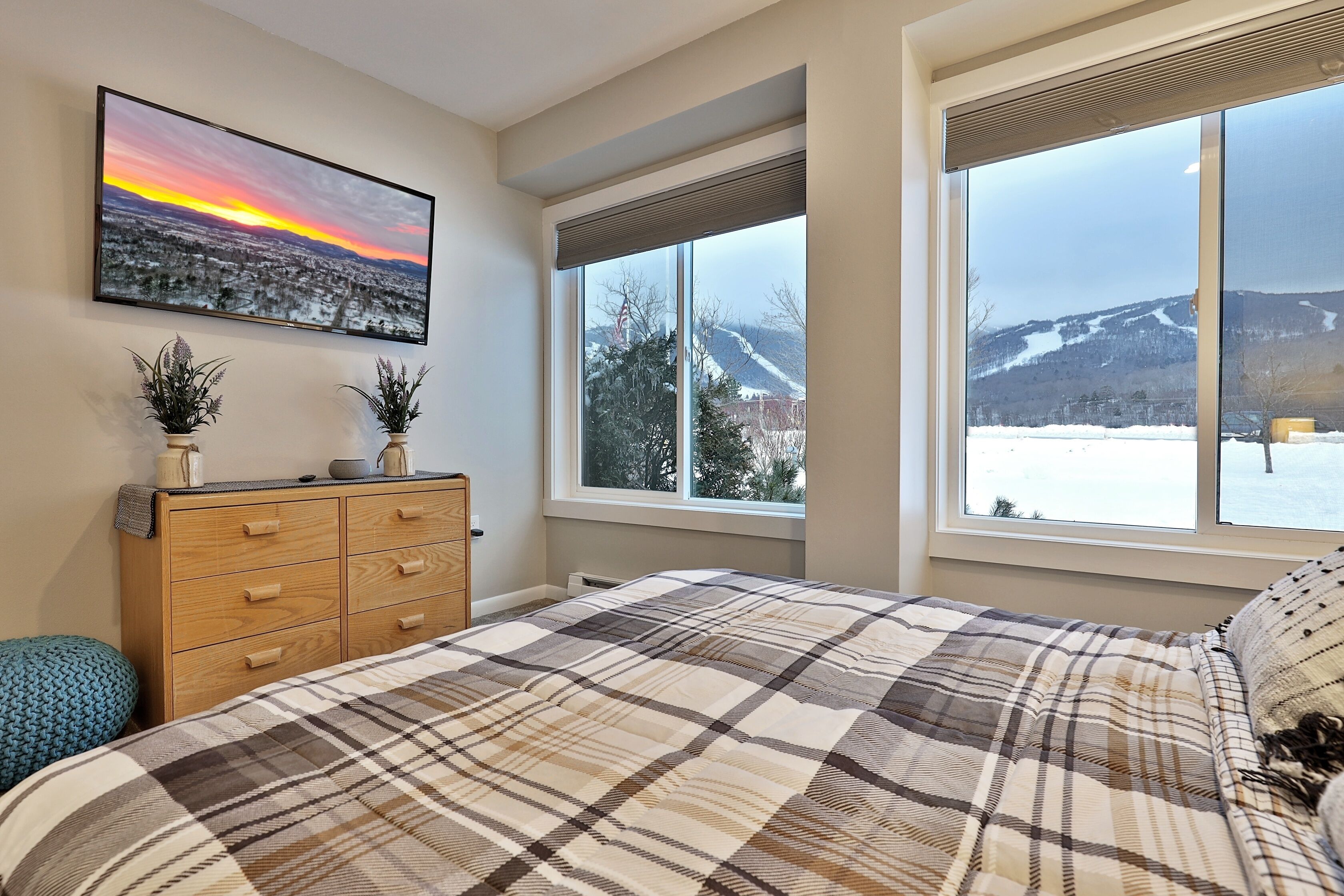 A bedroom with a large bed, a wooden dresser, two potted plants, and a cityscape painting.