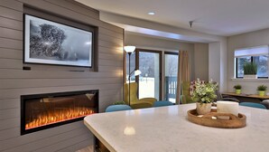 A modern living room with a fireplace, a large window, and a dining area with a marble table. - Mountain Green Resort by Killington VR - 2 Bedrooms (Killington)