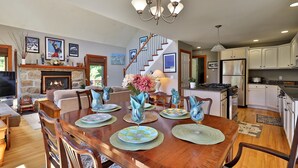 A spacious dining area with a wooden table set for four, surrounded by chairs. A chandelier hangs above, and the room features a stone fireplace and a staircase leading to another level. - Cortina Mountain Chalet - Outdoor Hot Tub - Close To Pico And Killington Mountains 3 Bedroom Home (Rutland)