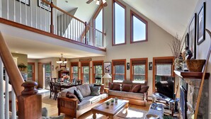 A spacious living area with a high ceiling, large windows, and a fireplace. - Cortina Mountain Chalet - Outdoor Hot Tub - Close To Pico And Killington Mountains 3 Bedroom Home (Rutland)