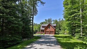 A house with a gravel driveway leading to a wooden garage, surrounded by dense green trees. - Cortina Mountain Chalet - Outdoor Hot Tub - Close To Pico And Killington Mountains 3 Bedroom Home (Rutland)