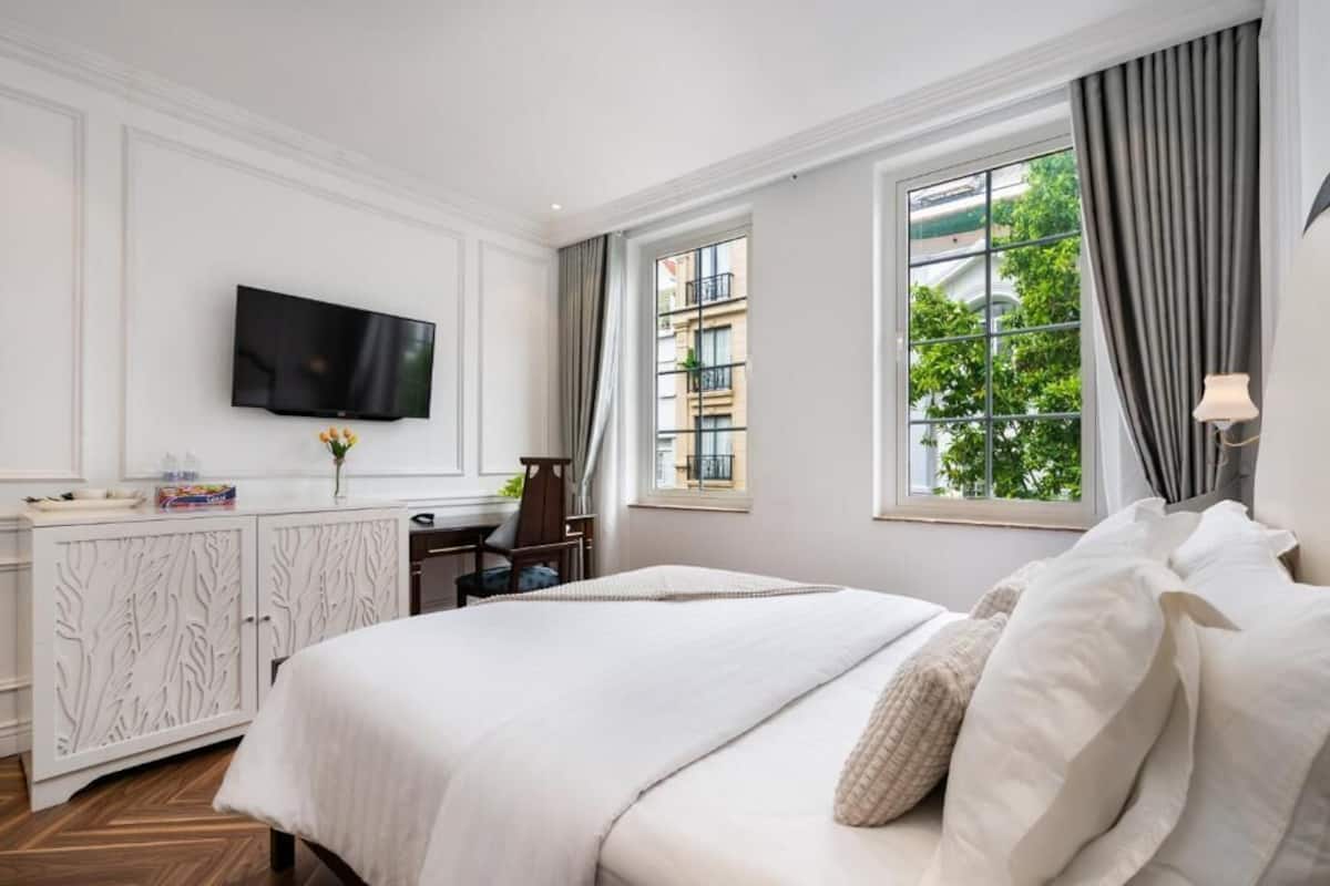 A hotel room with a large bed, a TV mounted on the wall, a desk with a chair, and a window with curtains.