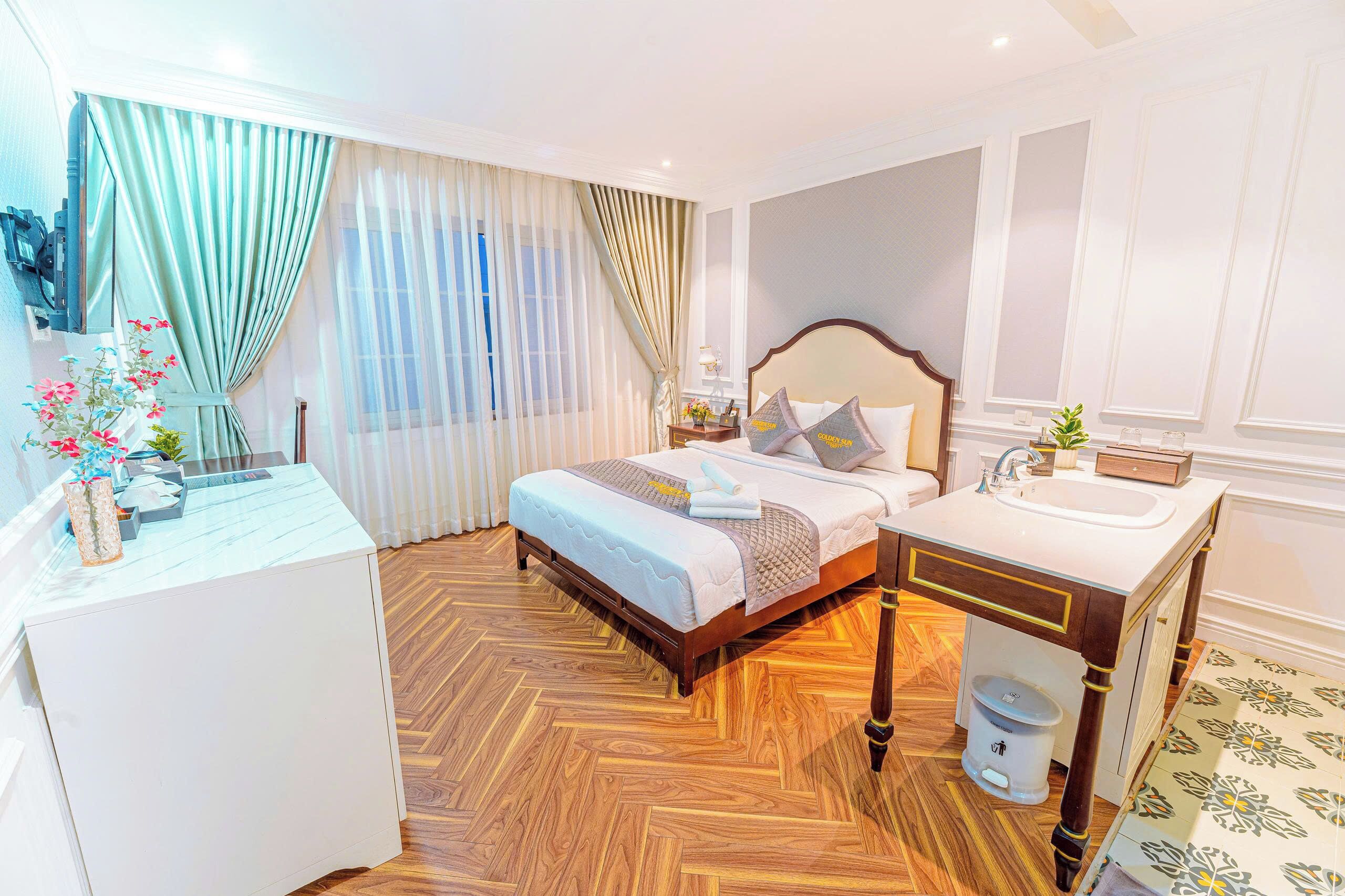 Habitación de hotel con suelo de madera en patrón de espiga, cama con cabecera blanca, mesita de noche con lámpara y baño con tocador y lavabo de madera.