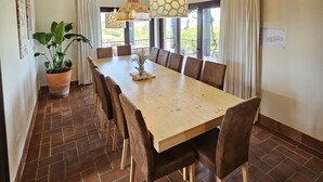 A dining area with a long wooden table, multiple chairs, hanging pendant lights, and a potted plant.