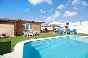 A pool area with a wooden building, white chairs, and blue lounge chairs.