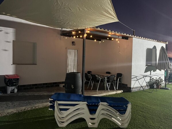 An outdoor patio with a table and chairs, a covered area with string lights, and a building with a window and air conditioning unit. - Holiday Home 'Villa Roulotte' with Shared Pool, Shared Garden and Wi-Fi (Chiclana de la frontera)