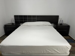 Ein Bett mit weißer Matratze und einem schwarzen Kopfteil, an beiden Seiten zwei schwarze Nachttische mit weißen Lampen.