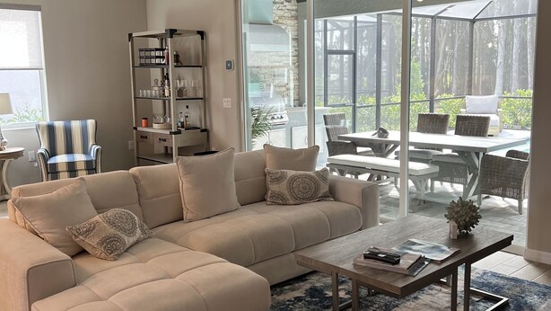 Un vaste salon comprenant un grand canapé d’angle beige, une table basse et un ventilateur de plafond. Une porte vitrée donne accès à un patio extérieur aménagé avec une table et des chaises.