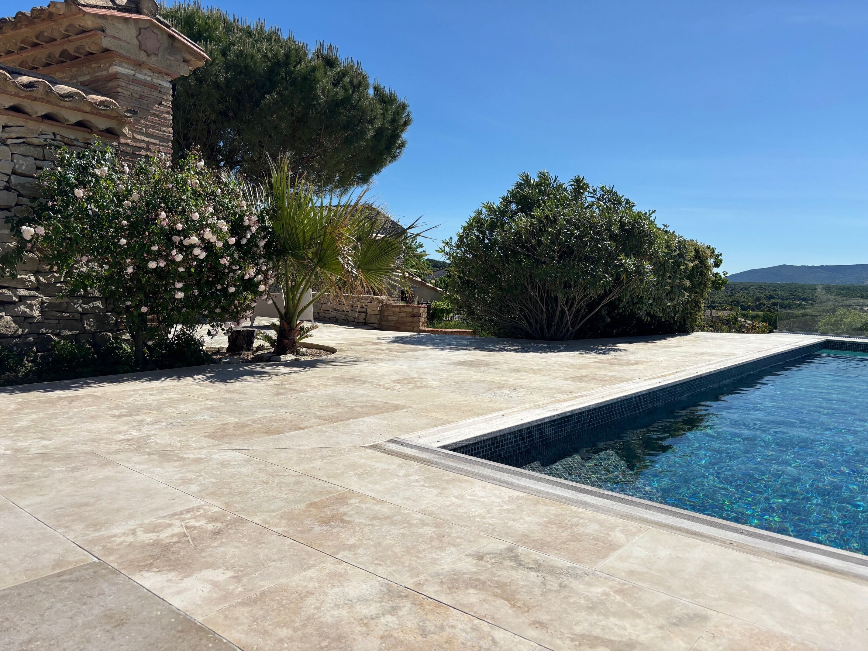 En swimmingpool med en flisebelagt terrasse, omgivet af træer og en stenvæg.