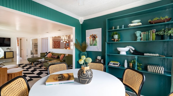 A modern living room with teal walls, a round dining table, wicker chairs, and a bookshelf filled with books and decorative items.