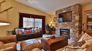 A cozy living room with a stone fireplace, leather furniture, and a television mounted on the wall. - The Tanglewood Lodge: Amazing Ski Home For Groups. Hot Tub. Close To Killington Resort! 4 Bedroom Home (Killington)