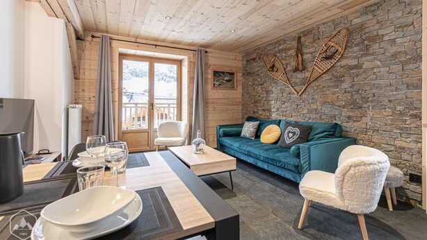 Ein gemütliches Wohnzimmer mit Holzbalkendecke, Steinwand, einem smaragdgrünen Sofa mit Kissen, einem weißen Sessel und einem Essbereich mit Tisch für zwei Personen.