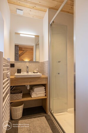 Un baño moderno con techo de madera, cabina de ducha con vidrio, lavamanos con espejo y toallas ordenadas en un estante.