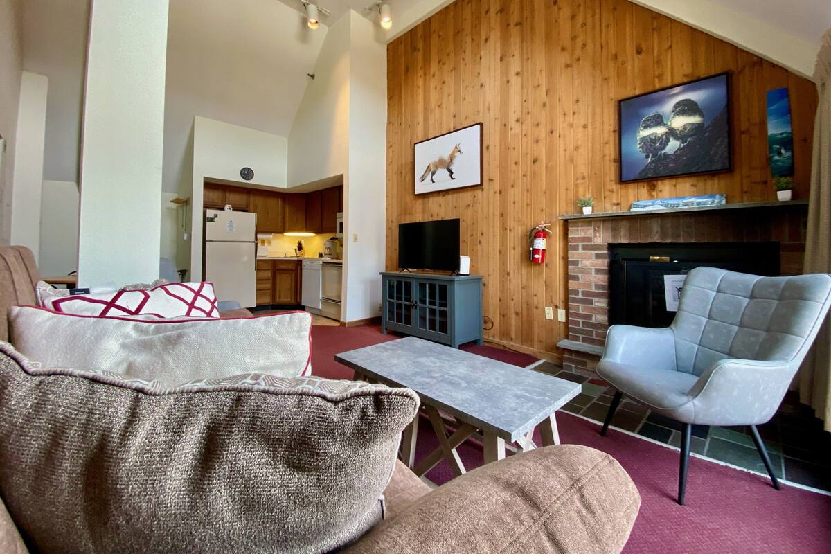Ein Wohnzimmer mit Kamin, Fernseher und einer Küche im Hintergrund.