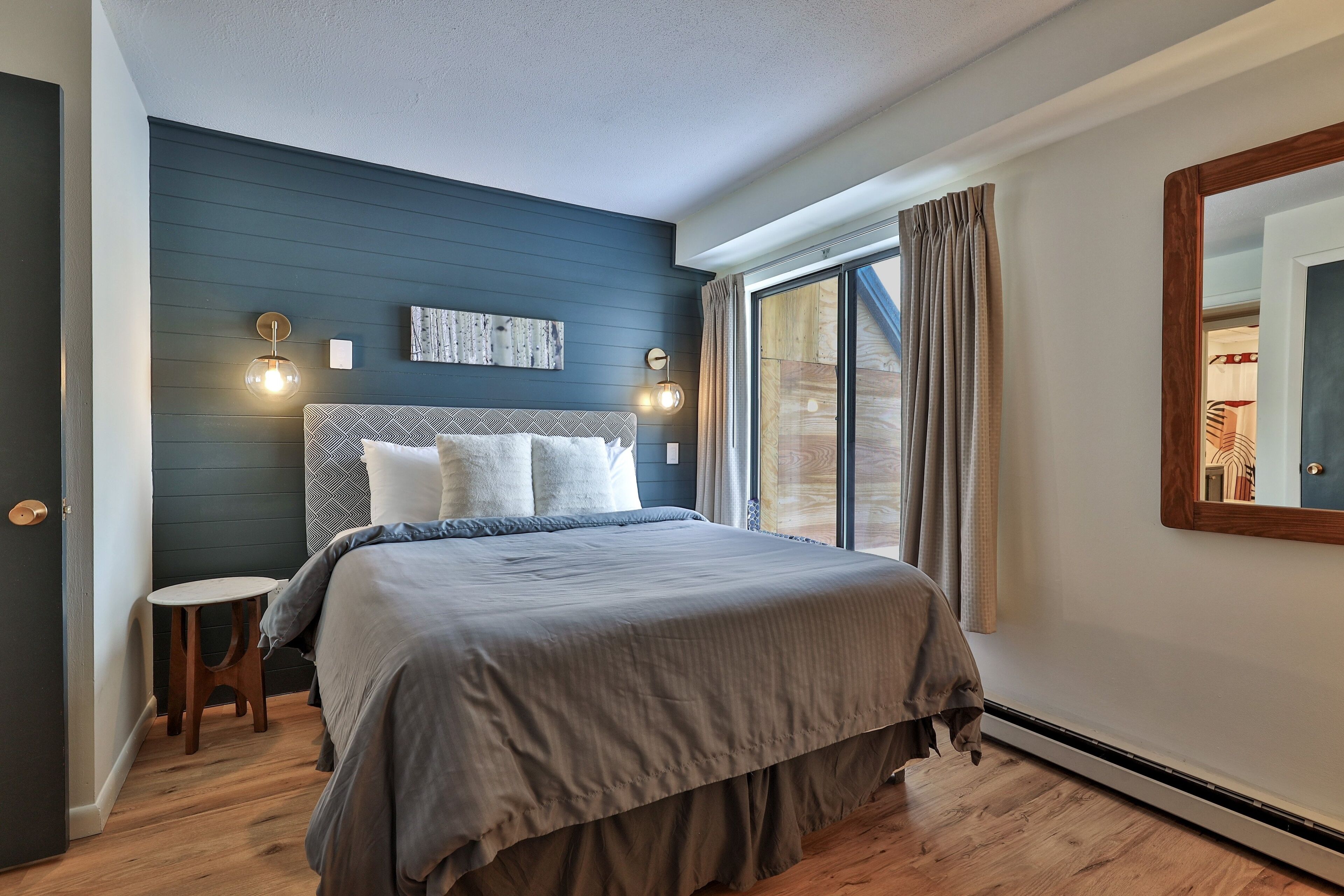 Une chambre à coucher comprenant un lit, une table de chevet, une lampe fixée au mur et une grande fenêtre avec des rideaux.