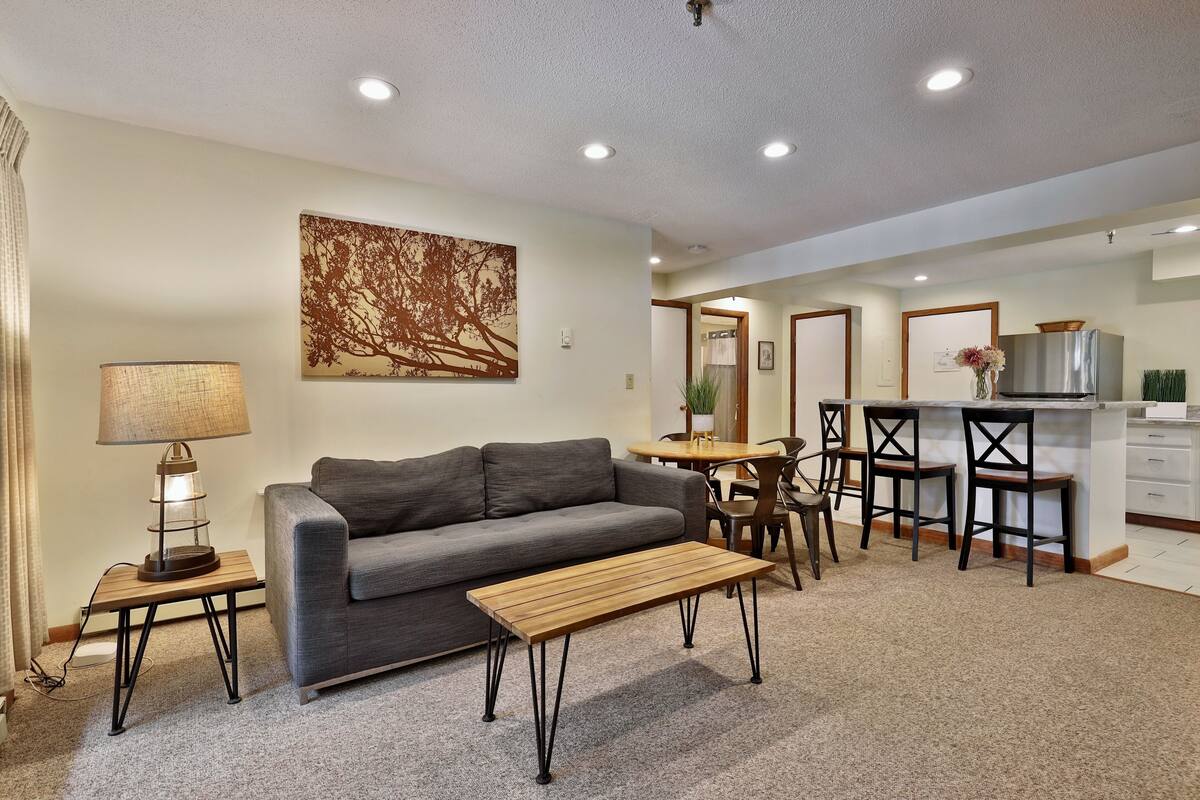 Ein modernes Wohnzimmer mit grauem Sofa, Couchtisch aus Holz und einer Lampe. Es gibt einen Essbereich mit Tisch und Stühlen. Der Raum hat eine neutrale Farbgestaltung und ein großes Kunstwerk mit einem Baum an der Wand.