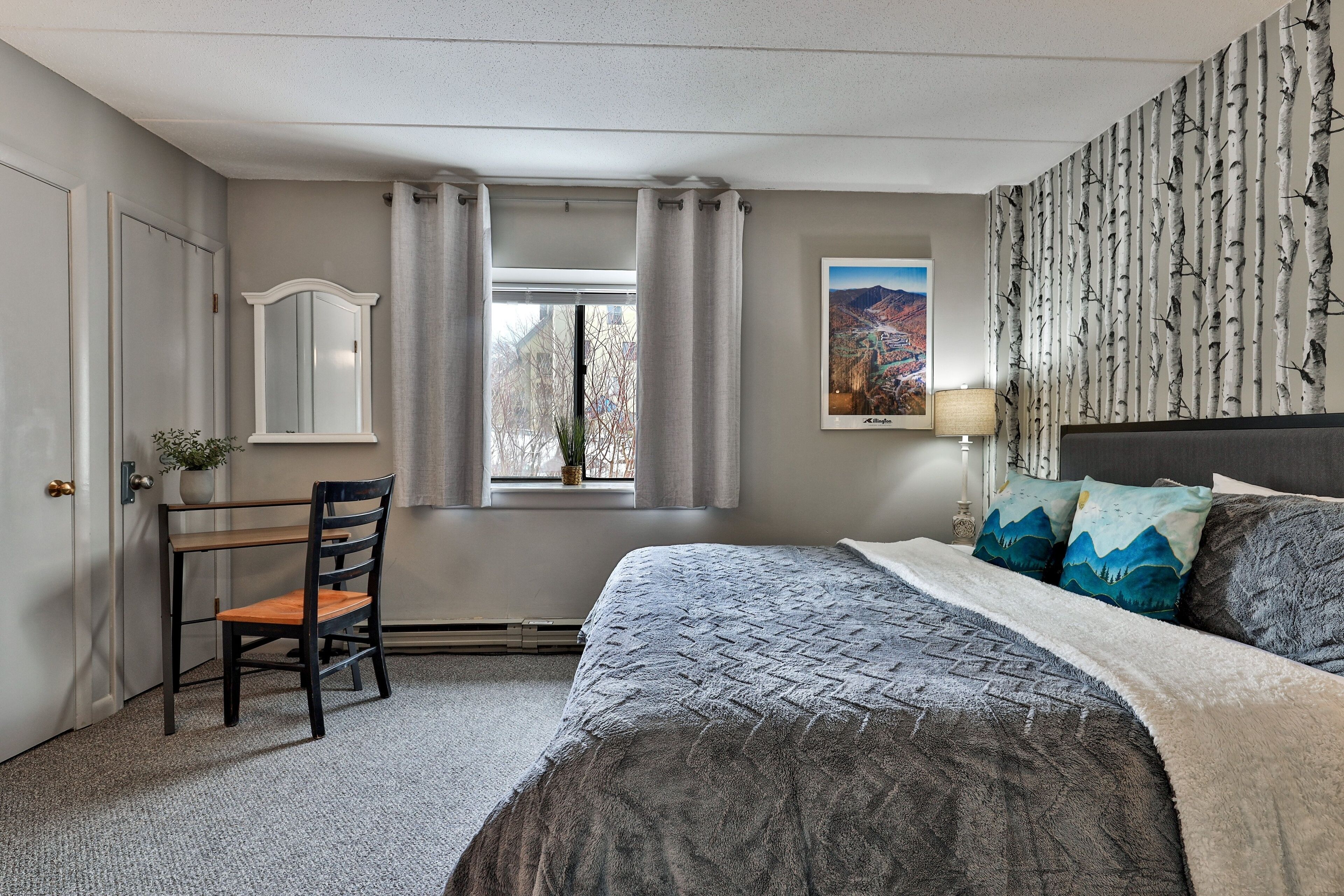 A bedroom with a bed, a desk, a chair, a window with curtains, a wall-mounted mirror, and a framed picture.