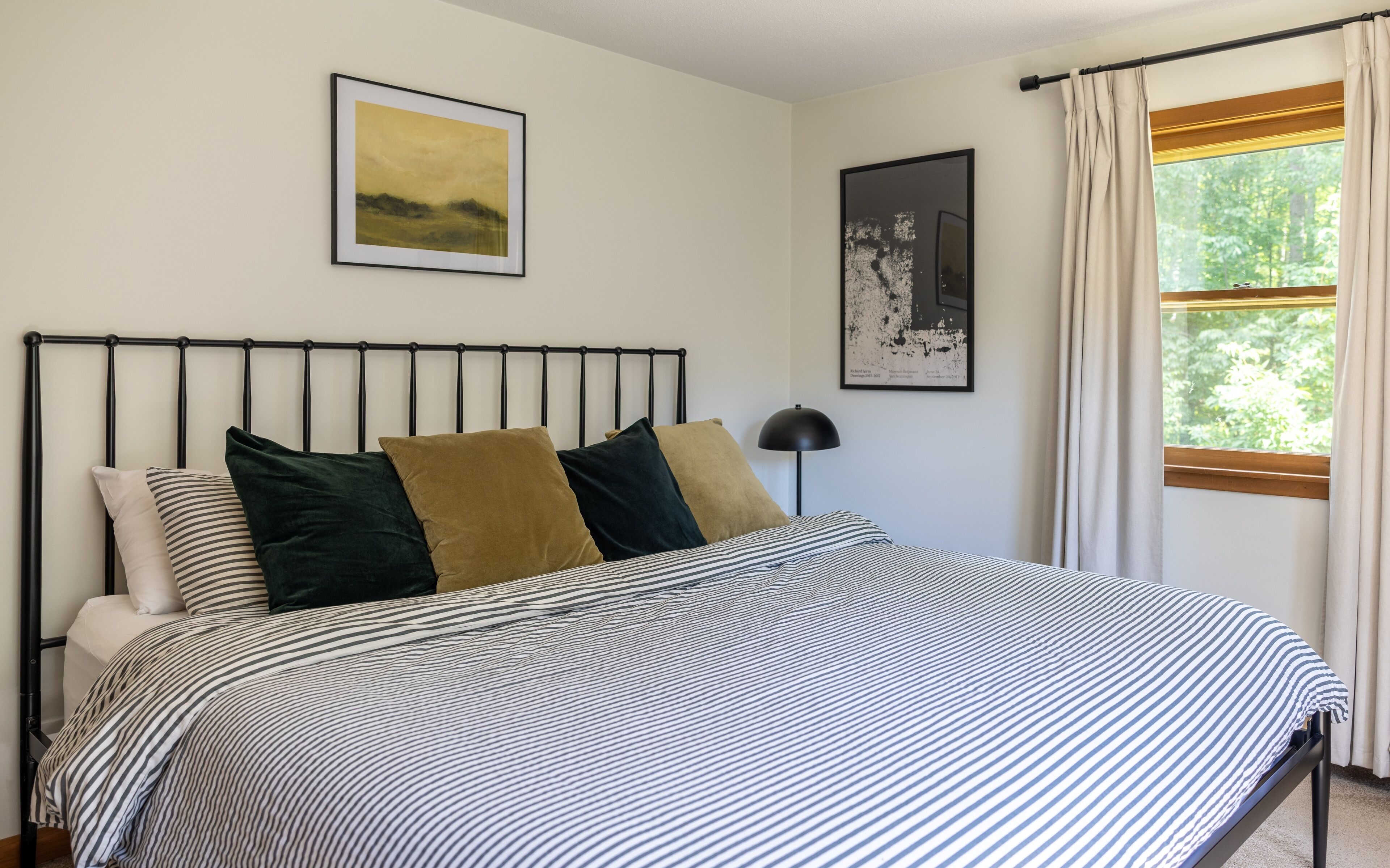 A bed with a striped bedspread, a metal headboard, and three pillows in different colors. There are two framed artworks on the wall, and a window with curtains.