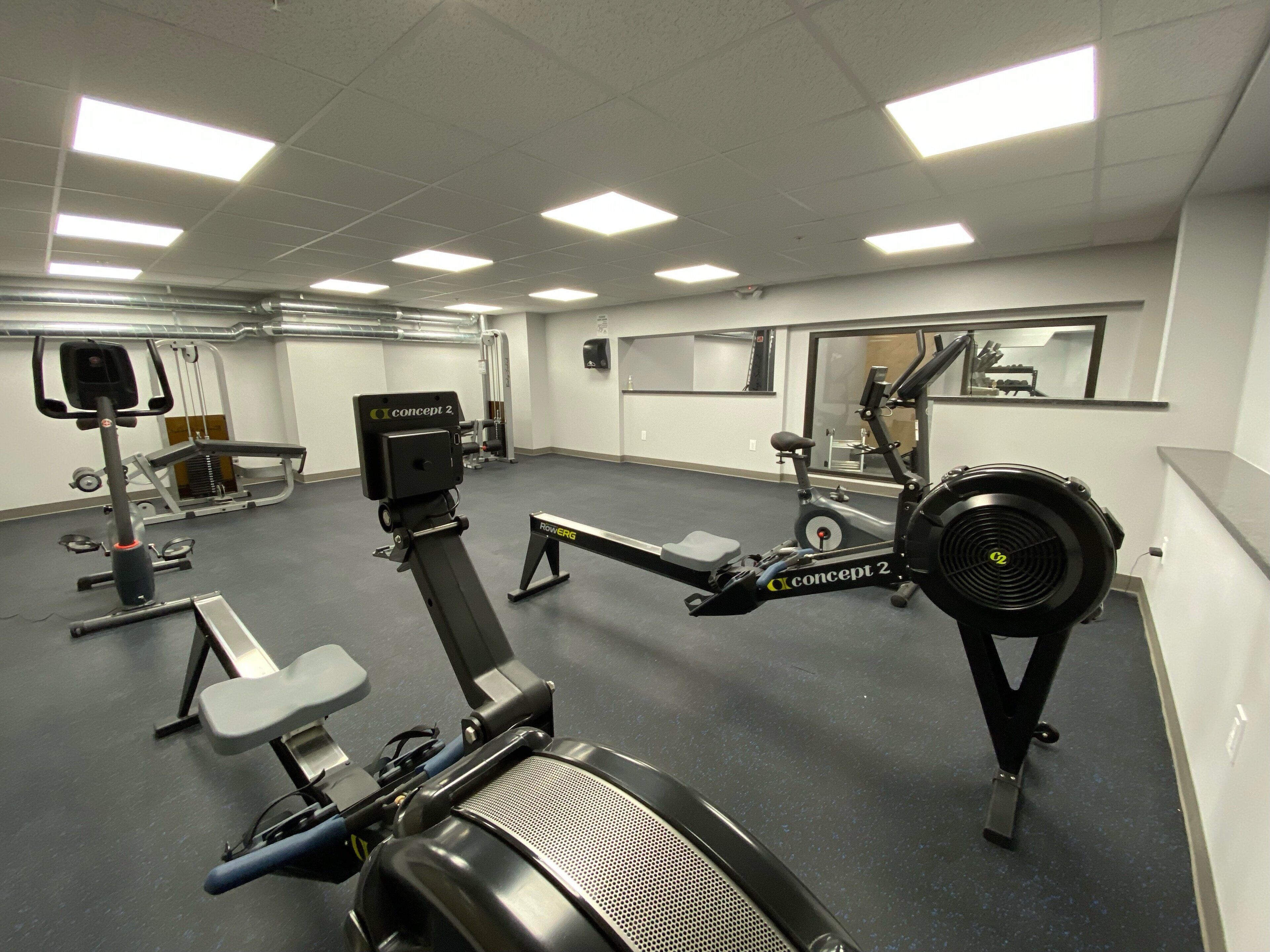 A gym with various exercise equipment, including a rowing machine and a stationary bike, with mirrors on the walls and fluorescent lighting.