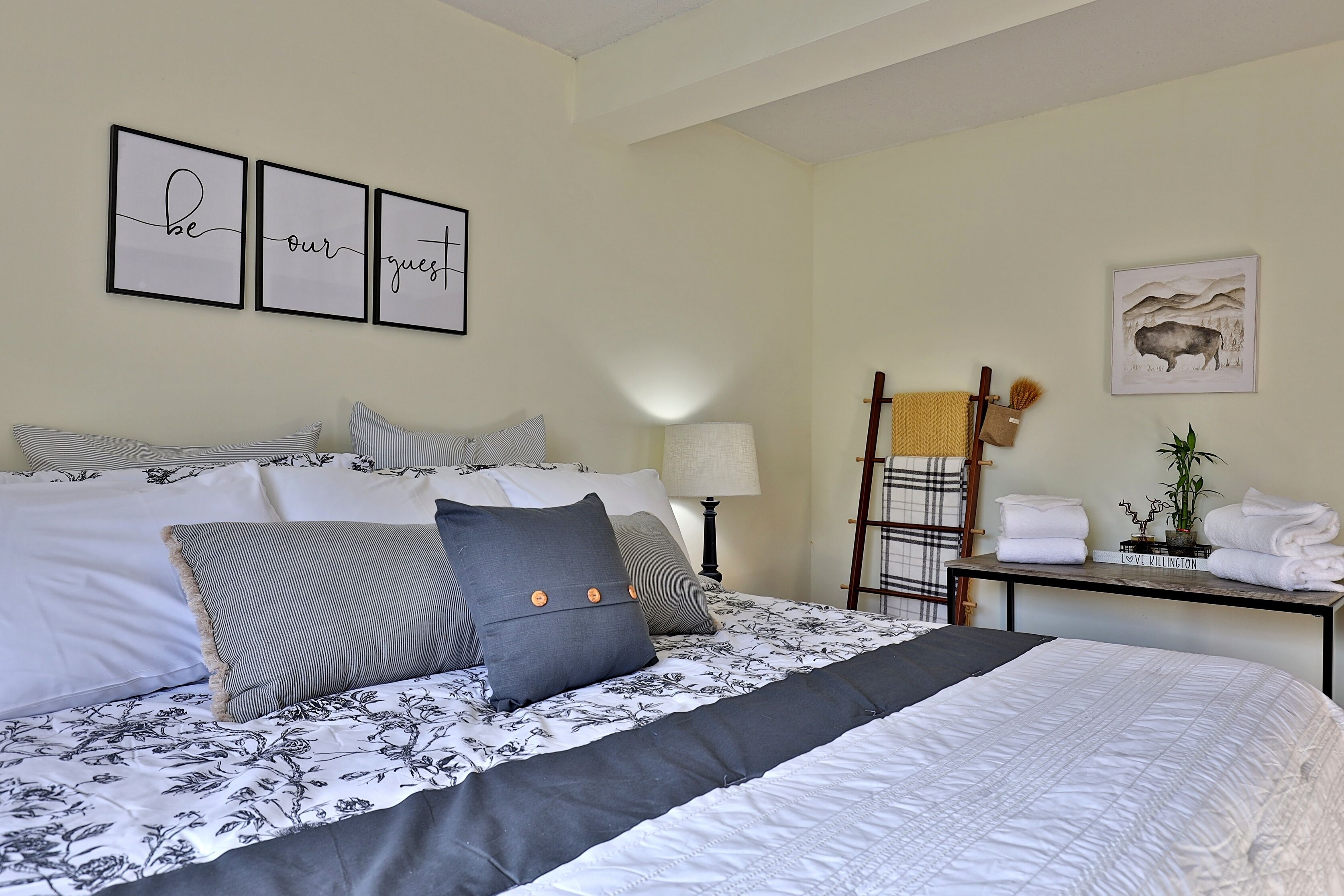 A neatly arranged bedroom with a large bed, a bedside table with towels, a lamp, and a framed picture on the wall.