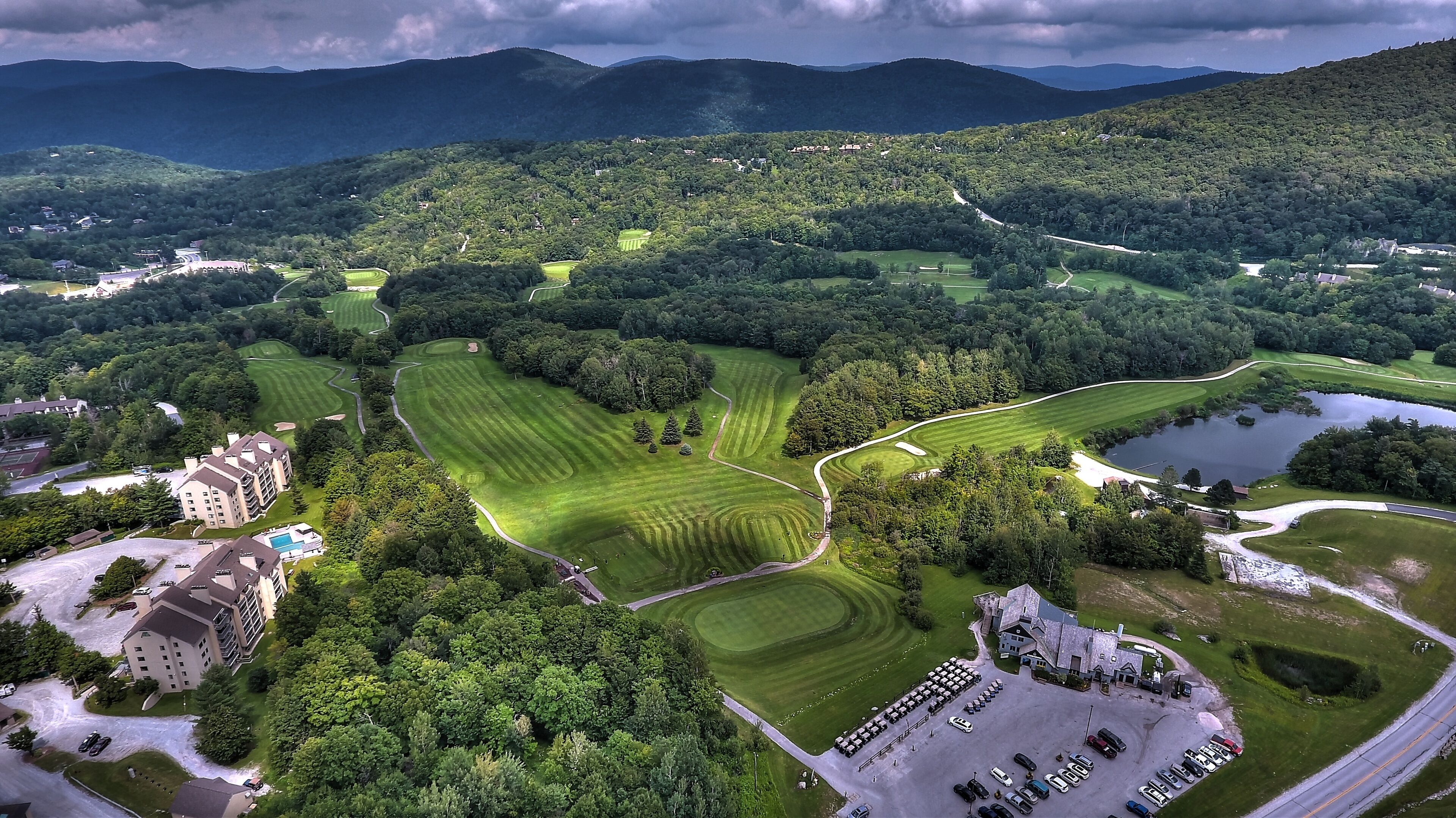 Vista aérea de un campo de golf con un club de campo, área de estacionamiento y colinas boscosas circundantes.