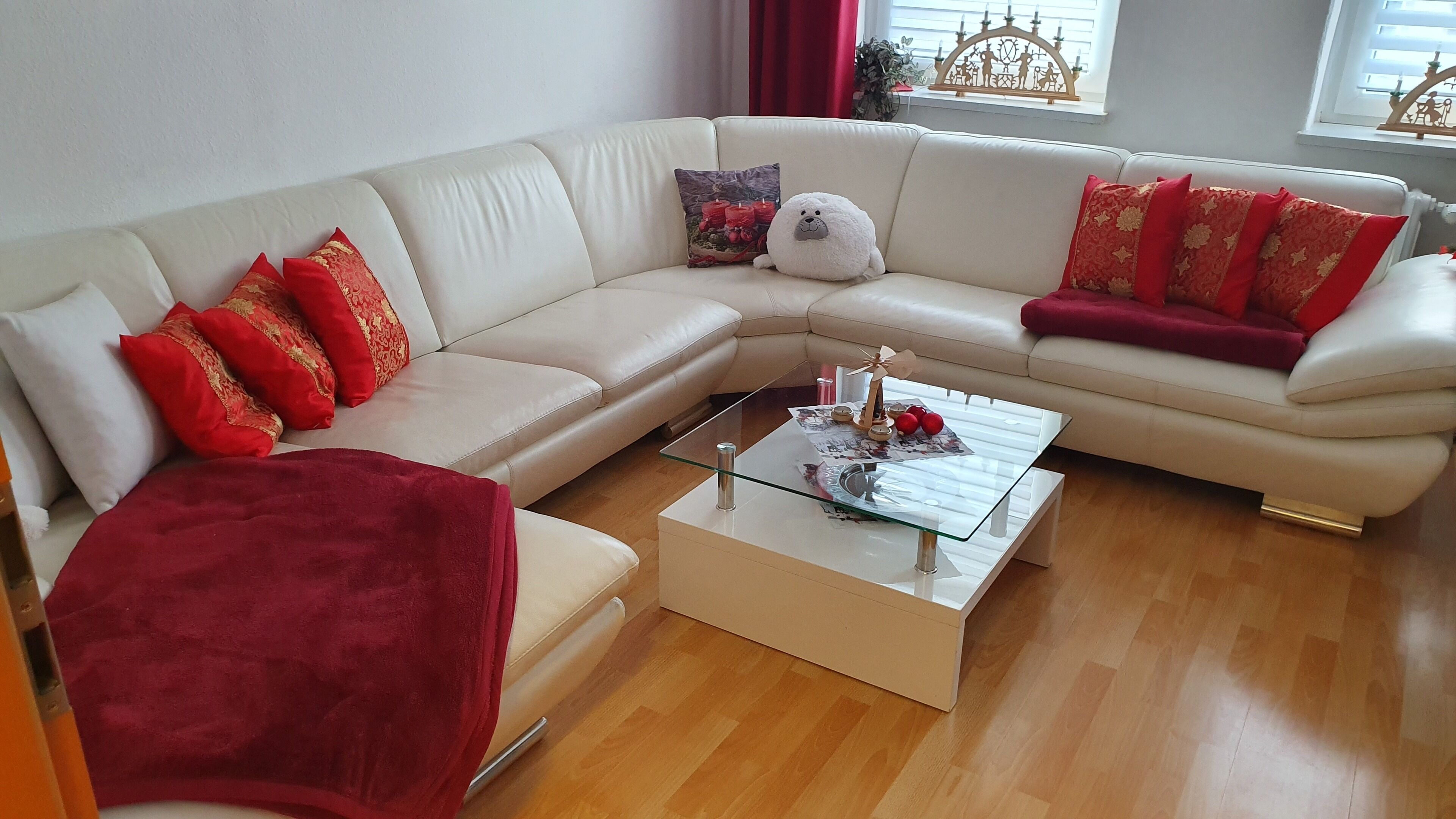 Ein modernes Wohnzimmer mit einem weißen Ecksofa, roten und goldenen Kissen, einem Glastisch und einer roten Tagesdecke.
