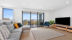 A modern living room with a grey sectional sofa, a flat-screen TV, and a balcony with a view of the city. - Wurrak Water View 20 Walk to Surf Shops & Cafes (Torquay)