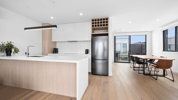 A modern kitchen with a central island, built-in appliances, and a dining area with a wooden table and chairs. - Wurrak Water View 20 Walk to Surf Shops & Cafes (Torquay)