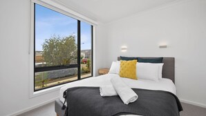 A modern bedroom with a large window, a bed with a grey headboard, white and yellow pillows, and a grey knitted throw. - Wurrak Water View 20 Walk to Surf Shops & Cafes (Torquay)