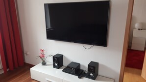 A modern living room with a flat-screen TV mounted on the wall, a white entertainment unit with speakers and a remote control, and a small vase with pink flowers on the unit. - Berg vacation apartment in Annaberg-Buchholz in the heart of the Ore Mountains  (Annaberg-Buchholz)
