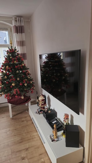 Ein Wohnzimmer mit Weihnachtsbaum, einem an der Wand befestigten Flachbildfernseher und einer weißen Unterhaltungsanlage mit diversen Gegenständen.
