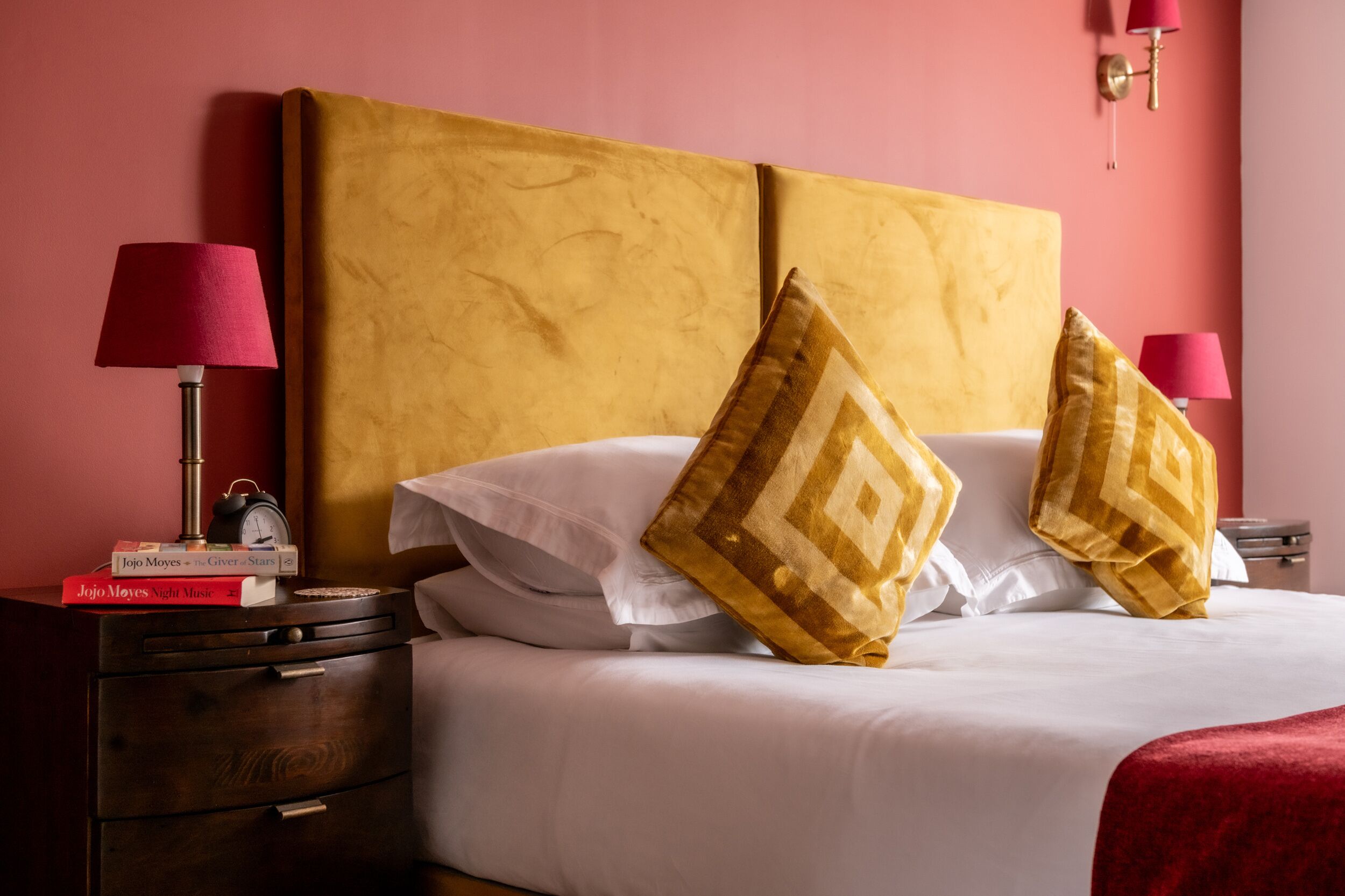 A neatly made bed with white linens and two decorative pillows, a bedside table with a lamp and a book, and a wall-mounted light fixture.
