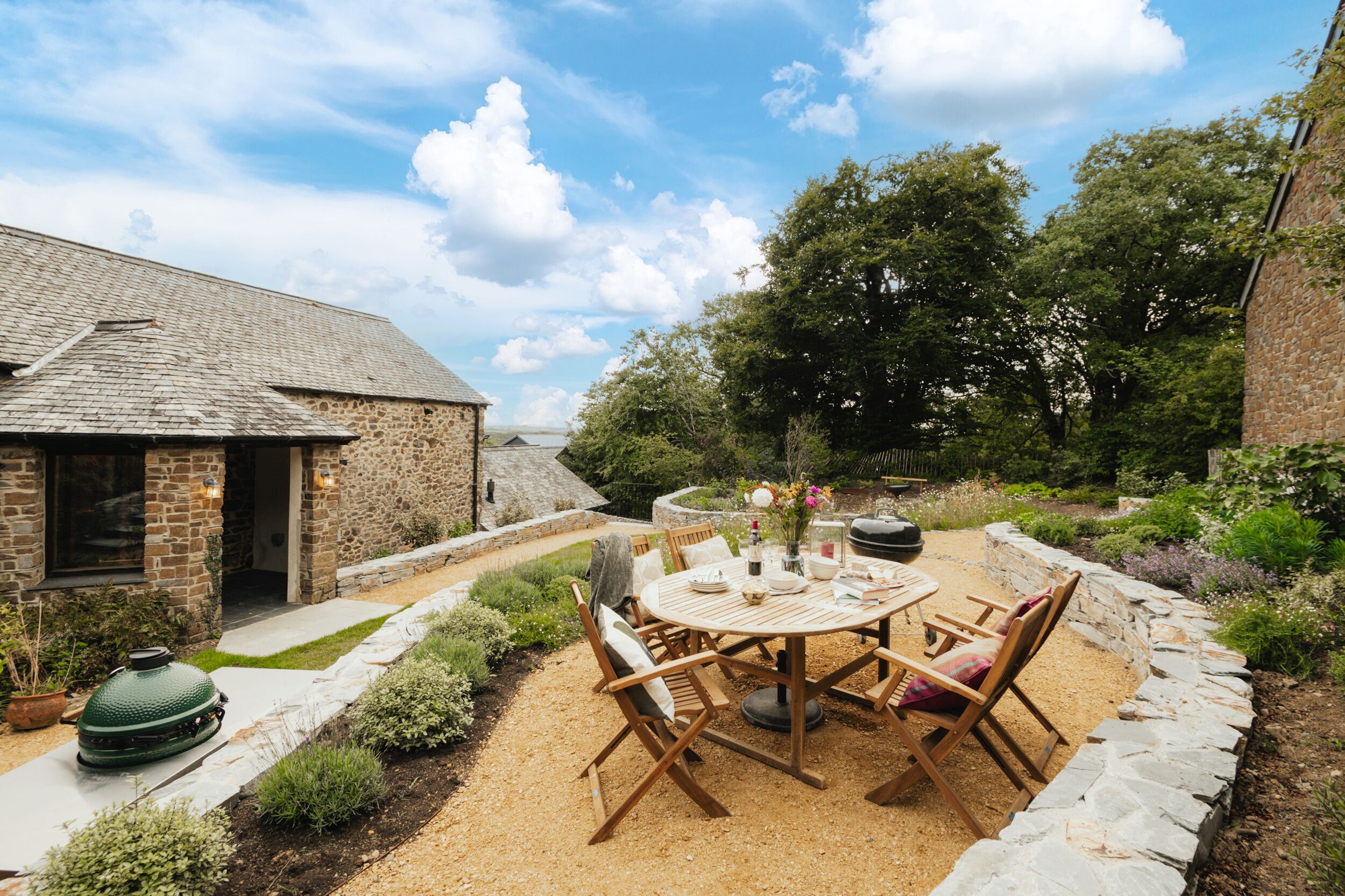 A patio with a round wooden table and chairs, surrounded by a stone wall and garden.
