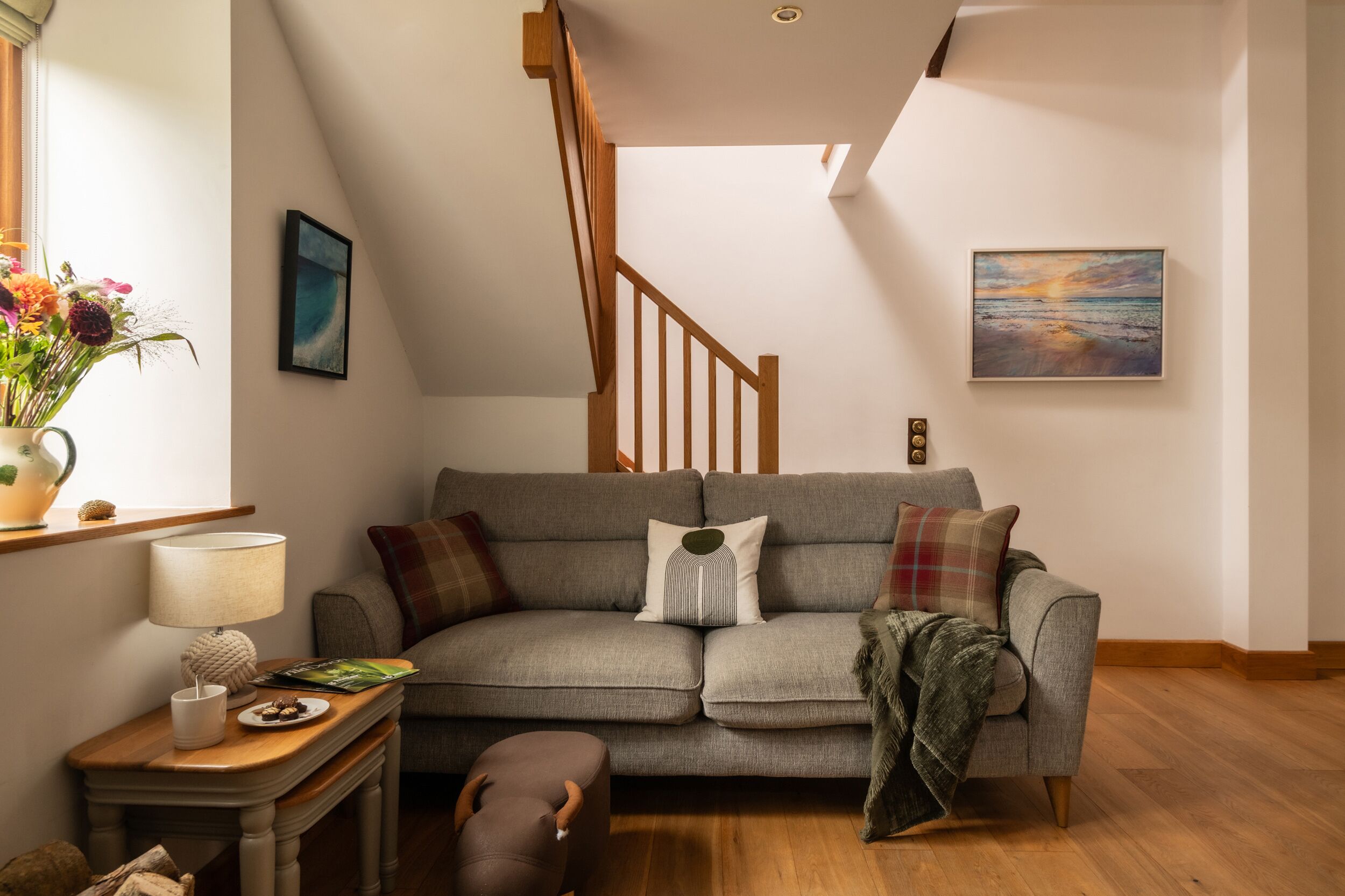 Un salon chaleureux avec un canapé gris, un escalier en bois et un tableau représentant un coucher de soleil.