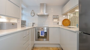 A modern kitchen with white cabinetry, stainless steel appliances, a built-in oven, and a wooden cutting board on the countertop. - Salty Sea Dog (Tintagel)
