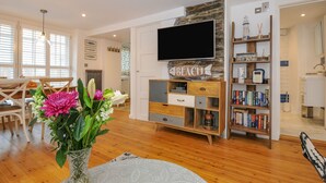 A spacious living room with a wooden floor, a flat-screen TV mounted on a stone wall, a bookshelf filled with books, and a dining area with a wooden table and chairs. - Salty Sea Dog (Tintagel)