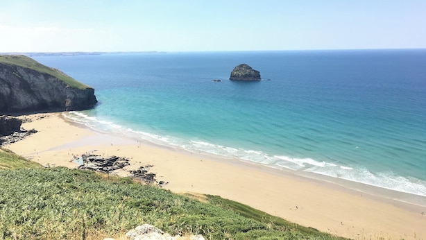 A coastal landscape with a sandy beach, a rocky outcrop, and calm sea. - Salty Sea Dog (Tintagel)