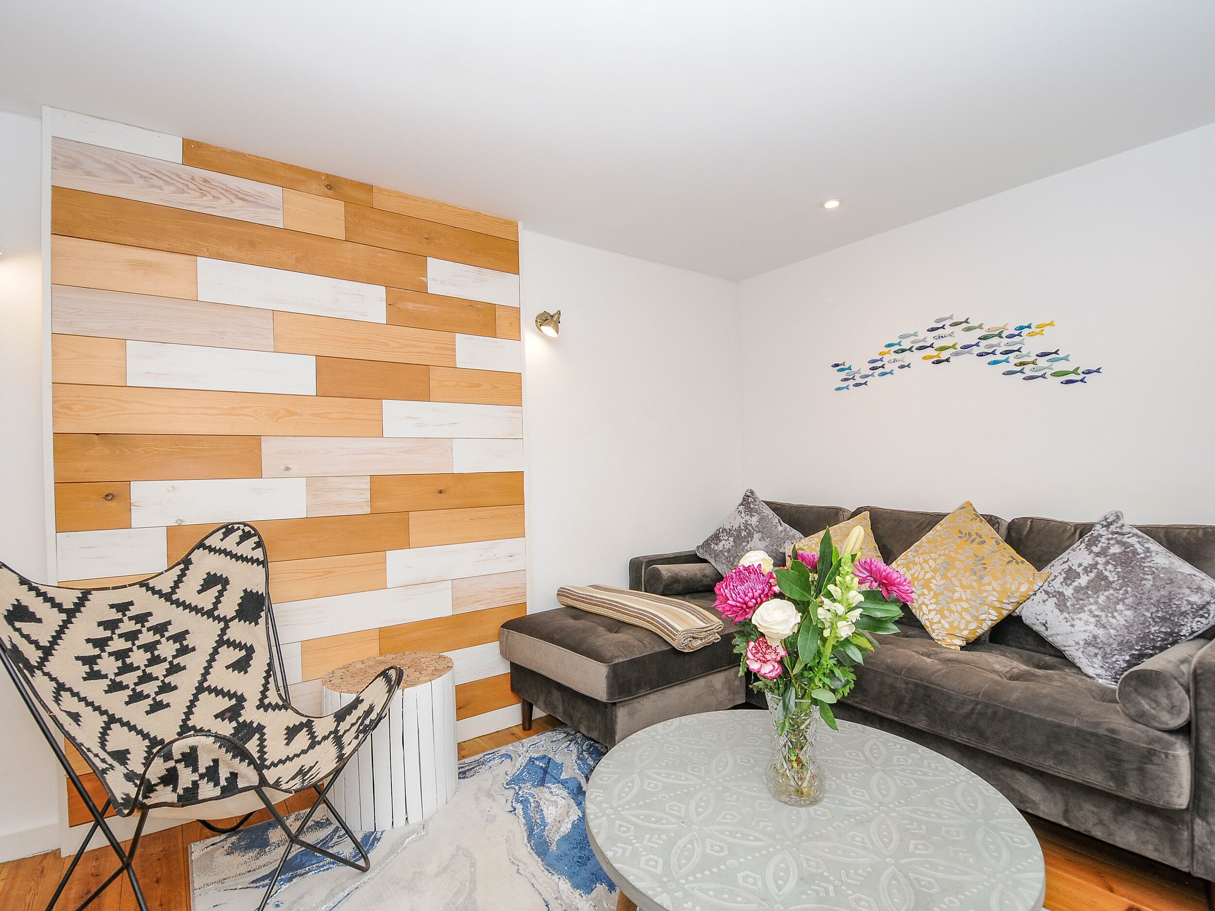 A modern living room with a wooden wall panel, a grey sofa with patterned cushions, a round coffee table with a vase of flowers, and a black and white patterned chair.