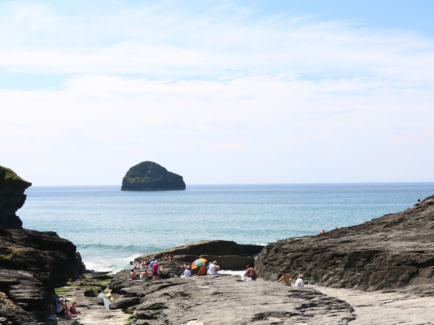 Une plage rocheuse où des gens se prélassent au soleil et se baignent, une grande formation rocheuse visible au loin, et une mer calme.