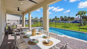 A covered patio with a pool, outdoor seating, and a view of a well-maintained lawn and pool area.