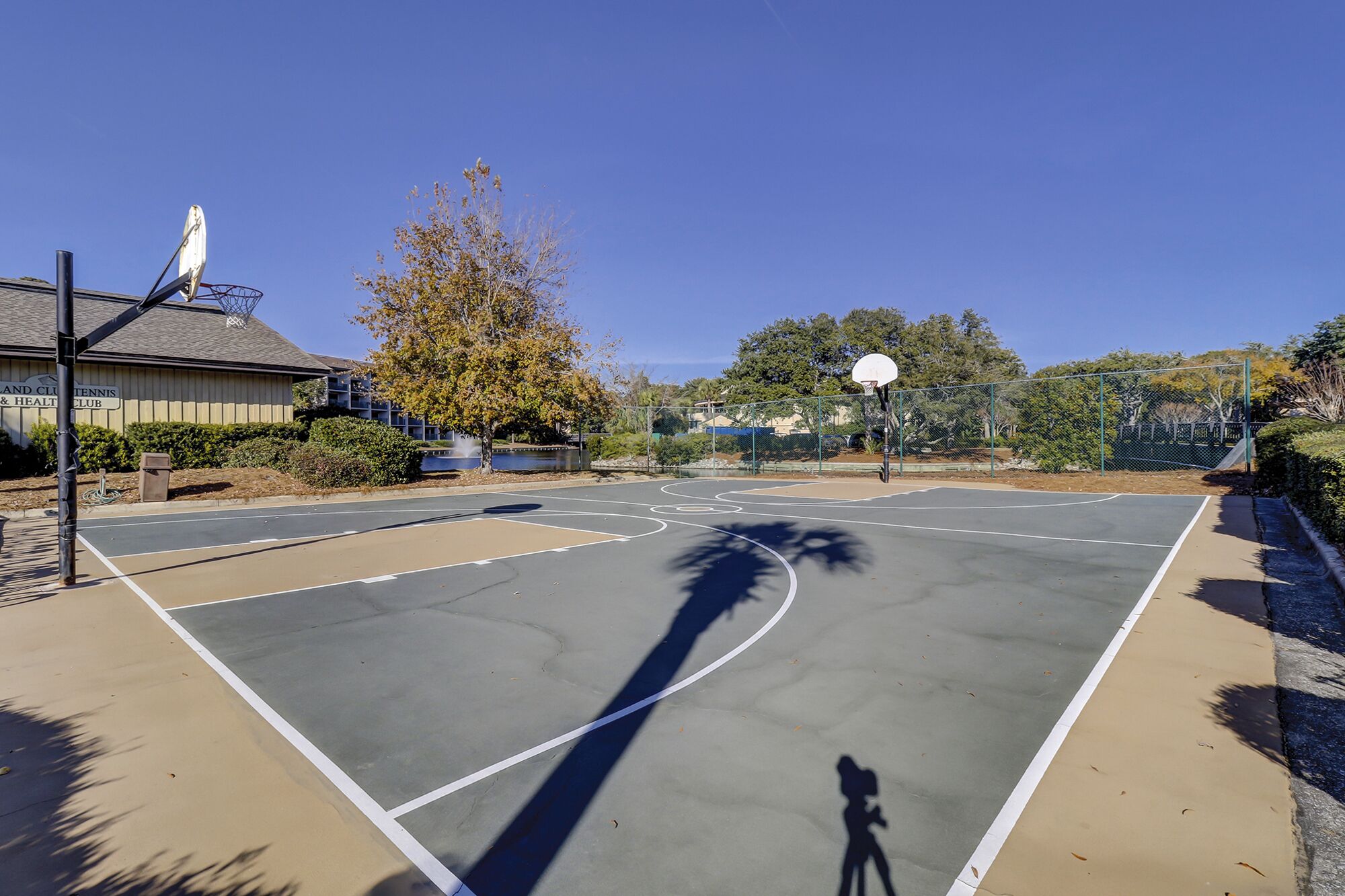 An outdoor basketball court with a clear sky, a tennis court building, and a tree in the background.
