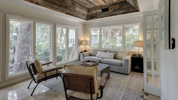 Un salon spacieux avec un plafond haut, de grandes fenêtres, un canapé gris, une table basse et une chaise en bois.