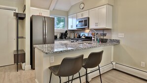 Una cocina moderna con armarios blancos, electrodomésticos de acero inoxidable, encimeras de granito y dos taburetes de bar.