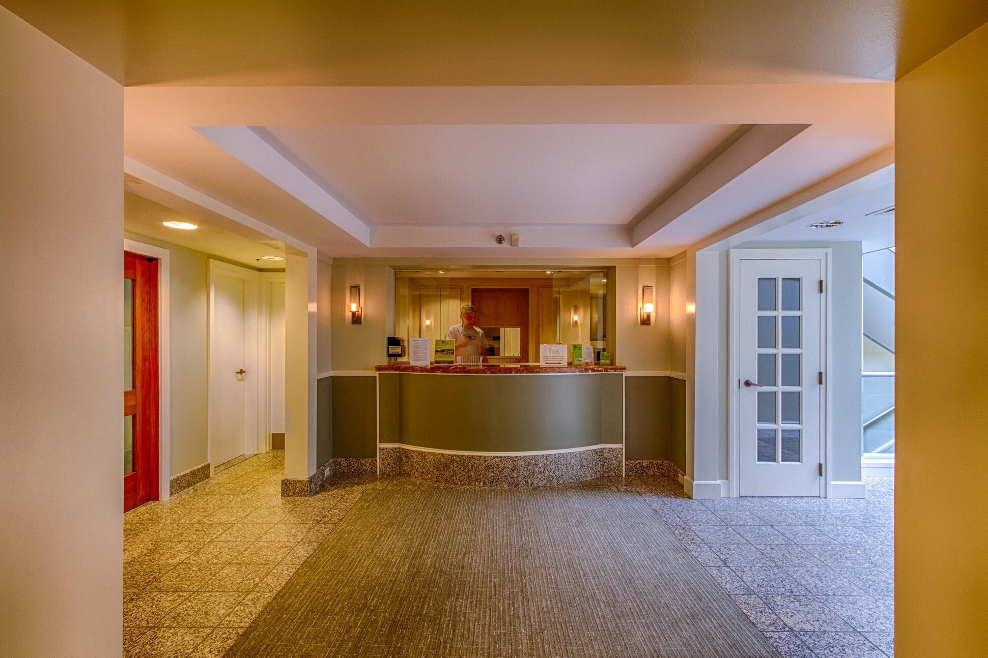 Un vestíbulo de hotel con una recepción, una puerta roja y una puerta de cristal que da a un balcón.