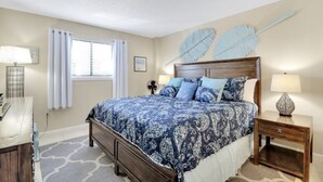 A bedroom with a large bed, a wooden nightstand, a desk, and a window with curtains. - Snow Lion On The Beach 2 Bedroom Villa by RedAwning (Hilton Head Island)