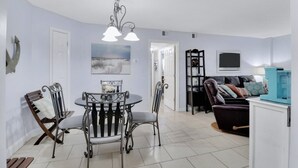 A dining area with a round table and chairs, a chandelier, and a view of the living room. - Snow Lion On The Beach 2 Bedroom Villa by RedAwning (Hilton Head Island)
