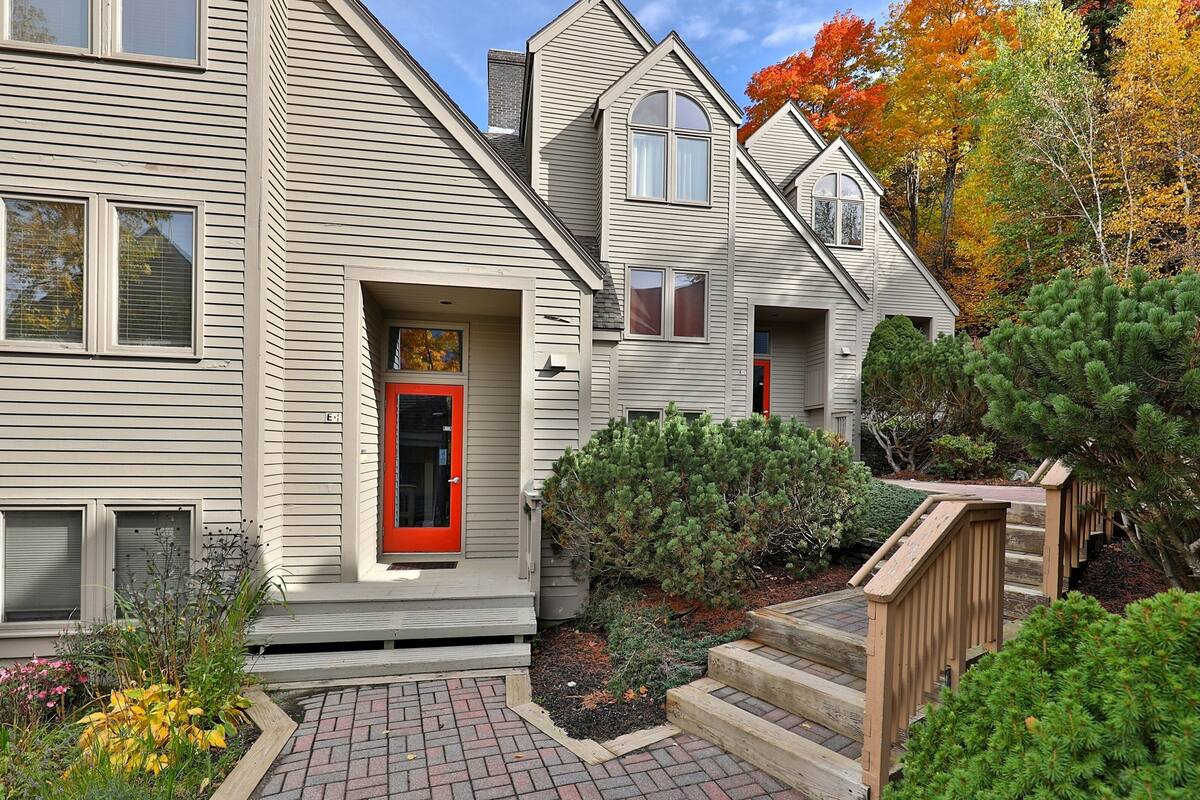 A two-story house with a red door, a brick pathway, and a wooden deck.