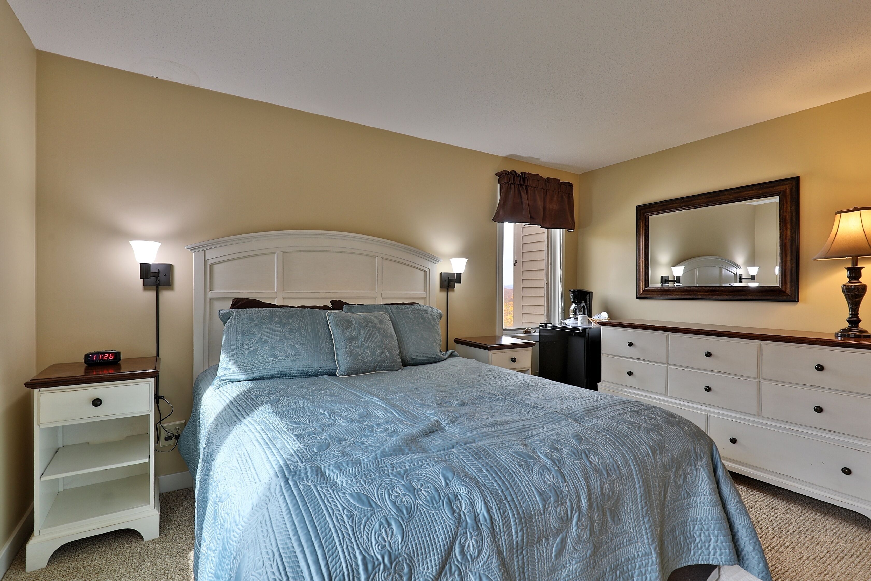 A bedroom with a bed, nightstands, a dresser, a mirror, and a window with curtains.