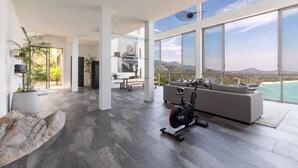 A spacious living area with a large window offering a scenic view, a modern exercise bike, and a minimalist interior design. - Villa Anushka – Luxury 6BR Koh Samui Villa with Staff, Breakfast & Sea Views (koh samui)