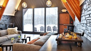 A cozy interior with a stone wall, a large window with a view of snow-covered trees, a wooden table with decorative items, and a sofa with a fluffy pillow. - Rent An Inn! Takeover The Birch Ridge W/ Your Group. Mountain Lodge: 11 Rooms, Bar, Fireplace. 28ppl 11 Bedroom Home (Killington)